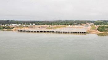 Un largo muelle de hormigón se extiende sobre un río. En tierra, hay edificaciones, maquinaria de construcción, postes de luz y vegetación con cielo nublado