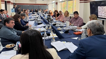 Un grupo de personas sentadas alrededor de una mesa larga y azul en una sala de reuniones. Hay documentos, vasos de agua y un micrófono visible, con ventanas al fondo