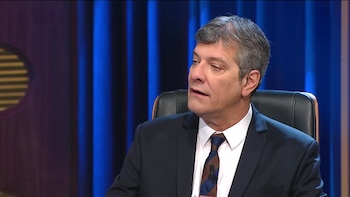Mario Pergolini con traje y corbata habla sentado en un sillón frente a una cortina azul