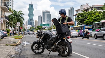 Un hombre con casco y chaleco reflectante abre una bolsa negra sobre una motocicleta junto a una vía con rascacielos y vehículos.