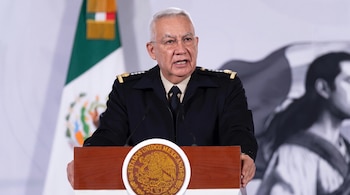 Un hombre con uniforme militar oscuro y gafas habla desde un podio con el escudo de México, una bandera mexicana y una figura borrosa al fondo.