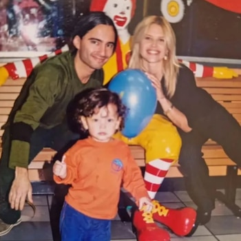 Un hombre de pelo oscuro, una mujer rubia y un niño de pelo rizado posan. Un globo azul. Detrás, una figura de Ronald McDonald y un banco