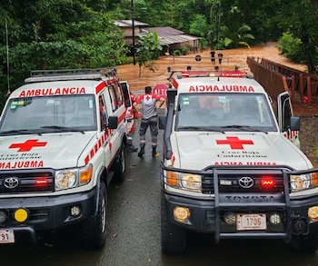 Tres rescatistas con uniformes de la Cruz Roja, cascos y chalecos, junto a un vehículo de rescate blanco. El fondo muestra un paisaje montañoso y húmedo con vegetación