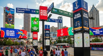 Calle de Atlanta con multitud y edificios, dominada por grandes carteles digitales y señales direccionales del Mundial 2026 hacia el Fan Zone y Mercedes-Benz Stadium.