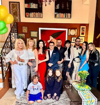 Un grupo de doce personas, incluyendo a Maribel Guardia, posa en una sala con globos, estanterías y cuadros. Un niño y dos niñas arrodillados en primer plano