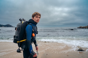 Un hombre con traje de buceo azul y negro, tanque de oxígeno y aletas, parado en la orilla de una playa. Detrás, el mar y un cielo nublado