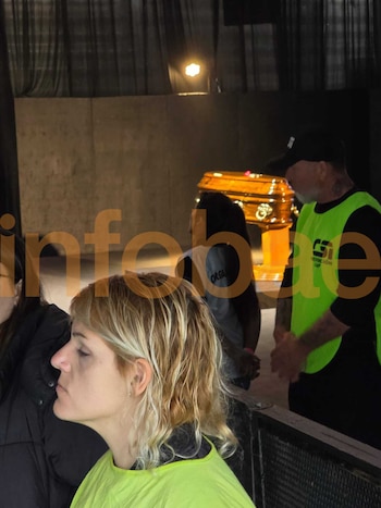 Vista de perfil de una mujer rubia en primer plano, con un piercing nasal, y en el fondo, un féretro dorado y un hombre de seguridad
