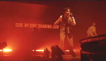 Hombre con chaleco y camisa blanca cantando en un micrófono sobre un escenario con iluminación roja intensa. Una pantalla proyecta texto y otra persona es visible al fondo