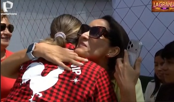 Pamela López abraza a una de sus hijas, quien lleva una camiseta a cuadros rojos con un gallo blanco. López usa gafas de sol. Varias personas observan