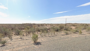 Amplia vista de un paisaje desértico con arbustos secos y un suelo pedregoso bajo un cielo azul claro. Un poste oscuro solitario se alza a la derecha