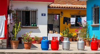 Seis tanques de gas doméstico (dos azules, dos rojos, dos plateados) en una acera frente a casas de colores vibrantes, con ropa tendida y plantas.