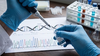 Manos con guantes azules utilizando una pipeta para transferir líquido a un vial sobre un documento con secuencia de ADN y equipo de laboratorio