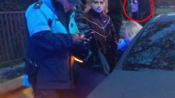 Vista nocturna de un agente de policía junto a varias personas y un coche, con una mujer de cabello oscuro destacada en un círculo rojo