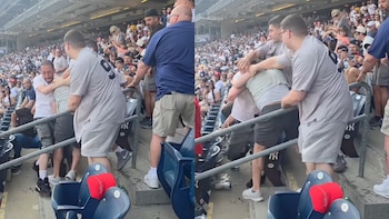 Varios hombres pelean en las gradas de un estadio de béisbol lleno de gente. Los asientos azules del estadio están desordenados, algunos volteados