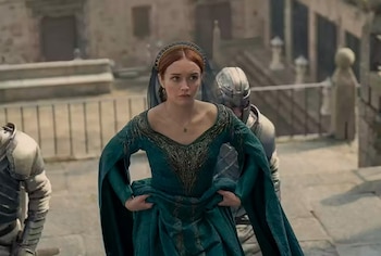 Una mujer con cabello rojizo, vestido largo verde y tocado dorado mira hacia adelante, flanqueada por dos figuras con armadura. De fondo, arquitectura de piedra y escalones