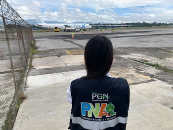 Espalda de una persona con chaleco de PGN y PNA mirando un avión Global X en la pista de un aeropuerto con valla de tela metálica