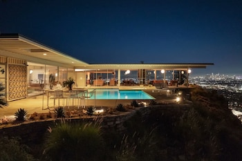 Casa de diseño moderno con ventanales de vidrio, piscina iluminada y muebles de terraza sobre una colina frente a luces nocturnas de la ciudad