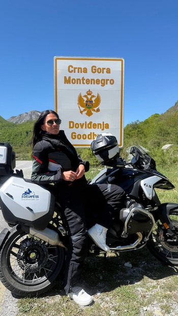 Carolina Baldini, con chaqueta y pantalón de moto negros y gafas de sol, posa apoyada en una motocicleta BMW blanca. Detrás, un cartel de Montenegro