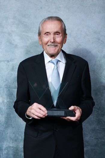 Hombre mayor, con traje oscuro, camisa blanca y corbata azul, sonríe a cámara mientras sostiene un trofeo con el logo 'WX' contra un fondo gris