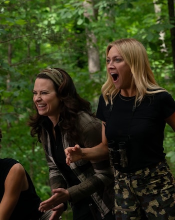 Malin Akerman con boca abierta de asombro y Brittany Snow riendo fuertemente en un bosque. Akerman lleva una camiseta negra y pantalones de camuflaje
