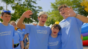 Cuatro jóvenes sonrientes con camisetas azules 'Walking 4 Friendship' saludan a la cámara en un evento al aire libre con árboles y un castillo inflable al fondo