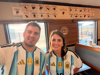 Un hombre y una mujer sonrientes con camisetas de fútbol de Argentina en un restaurante con decoración de mapas provinciales argentinos y el logo 'Los Hornos'