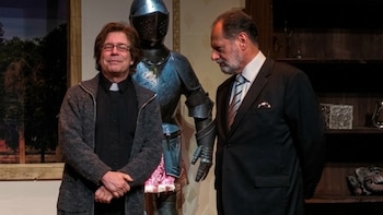 Dos hombres posan en un escenario, uno con cuello clerical y el otro con traje. Una armadura de metal azul se ubica entre ellos. Fondo con árboles y una estantería oscura
