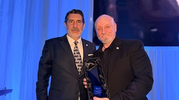 Dos hombres posan en un escenario iluminado de azul. Uno, con traje y corbata, entrega un premio a un hombre calvo con barba, vestido de negro