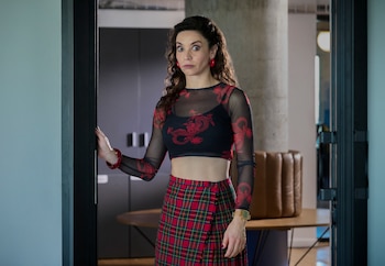 Mujer con cabello rizado, top de malla con dragones rojos, falda escocesa roja y verde, pendientes y pulsera rojos, reloj, abdomen descubierto, en una puerta
