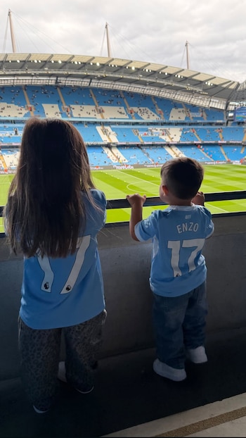 Dos niños vistos de espaldas con camisetas celestes con el número 17 miran el campo desde la baranda de las tribunas de un estadio