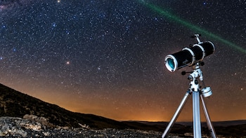 Telescopio negro sobre trípode en un terreno rocoso frente a un cielo nocturno con estrellas y una línea de luz verde