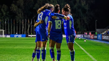 Cuatro jugadoras de fútbol con uniforme azul se agrupan en el campo de juego de un estadio por la noche. Se ven los números 13 y 18 en sus espaldas.