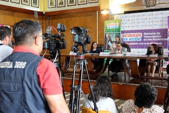 Camarógrafo de Prensa IGSS filma conferencia de prensa sobre vacunación con ponentes en mesa y pancartas de '¡VACUNACIÓN EN ACCIÓN!' de fondo
