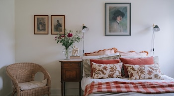 Um quarto com cama, travesseiros, cobertor justo, armário, vaso de flores, estátua, livro. Uma cadeira de vime, duas luminárias e quatro quadros na parede.