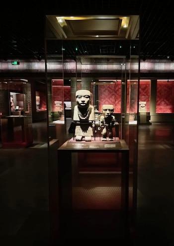Dos figuras de piedra prehispánicas, una grande y otra pequeña, se exponen en una vitrina de cristal iluminada en una sala de museo oscura con paredes rojas