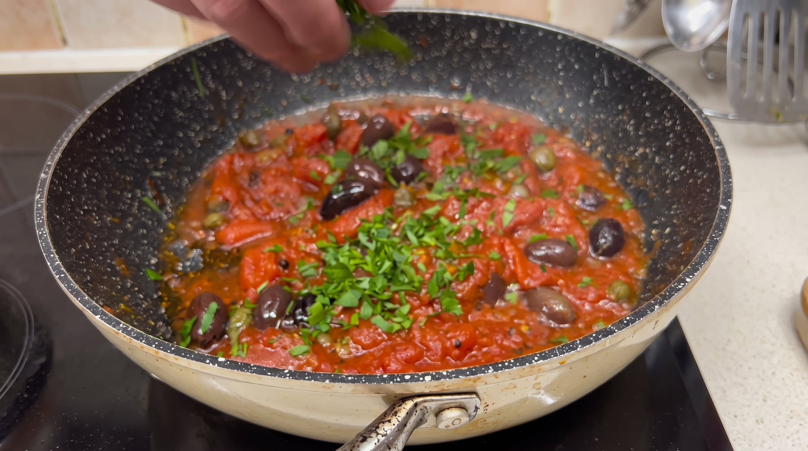 Una mano esparce hierbas frescas picadas sobre una sartén con salsa de tomate, aceitunas negras y alcaparras, cocinándose en una estufa. (@rafuel55/Instagram)