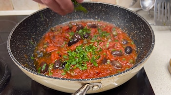 Una mano esparce hierbas verdes picadas sobre una sartén con salsa roja de tomate, aceitunas negras y alcaparras en una estufa.
