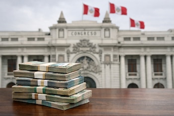 Pila de billetes de soles peruanos sobre una mesa de madera con el edificio del Congreso de la República del Perú y banderas al fondo.