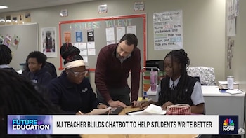 El profesor Scott Kern se inclina sobre un escritorio, interactuando con dos estudiantes sentadas que miran un portátil y documentos en un aula escolar