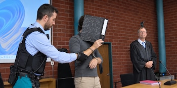 Un agente sujeta a un hombre esposado que cubre su cara con una carpeta negra frente a una persona con toga en una sala judicial