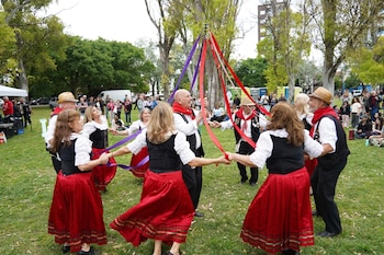 Seis personas en trajes folclóricos con faldas rojas y chalecos negros bailan alrededor de un palo de mayo con cintas de colores en un parque