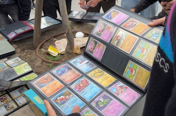 Manos de una persona sosteniendo un álbum negro abierto con filas de cartas Pokémon de colores brillantes. Otros álbumes y el suelo de tierra son visibles