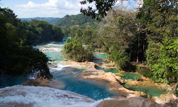 Una vista aérea de cascadas con múltiples pozas de agua turquesa y formaciones rocosas de color ocre, rodeadas por una densa selva verde
