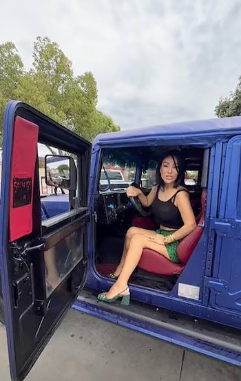 Una mujer joven está sentada al volante de un gran vehículo azul con la puerta del conductor abierta. Viste un top negro y una falda a cuadros verdes