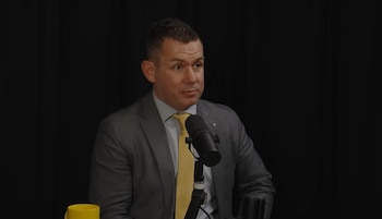 James Betts, un hombre con traje gris y corbata amarilla, habla frente a un micrófono negro. Fondo oscuro y una taza amarilla a la izquierda