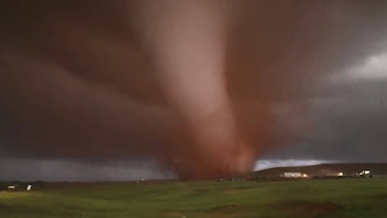 Un gran tornado de color marrón rojizo se extiende desde un cielo oscuro hacia un campo verde, con algunas estructuras iluminadas en la distancia