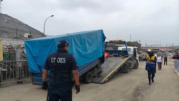 Un agente de seguridad de espalda observa una grúa azul que carga un furgón cubierto con una lona azul en una calle de tierra