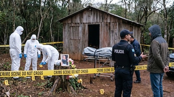 Equipo forense en trajes blancos trabaja tras cinta amarilla en escena de crimen rural, junto a una cabaña de madera. Un oficial y un hombre esposado observan.