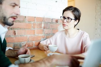 Una mujer con gafas y pelo corto muestra una cara fruncida mientras mira al hombre sentado junto a ella en una cafetería, que mira hacia un lado hablando con otra persona fuera de plano. Sobre la mesa, cada uno tiene una taza de café frente a él