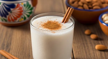 Primer plano de un vaso de horchata fría con canela en polvo y dos ramas de canela, sobre mesa de madera, con almendras y recipiente artesanal al fondo.